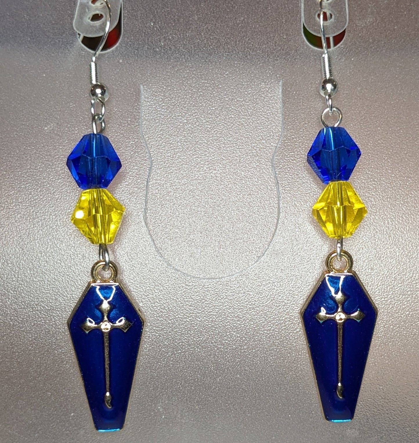 A pair of Vamp Goth Earrings featuring coffin-shaped charms in enameled in blue with a gold cross with faceted blue and yellow bead accents.