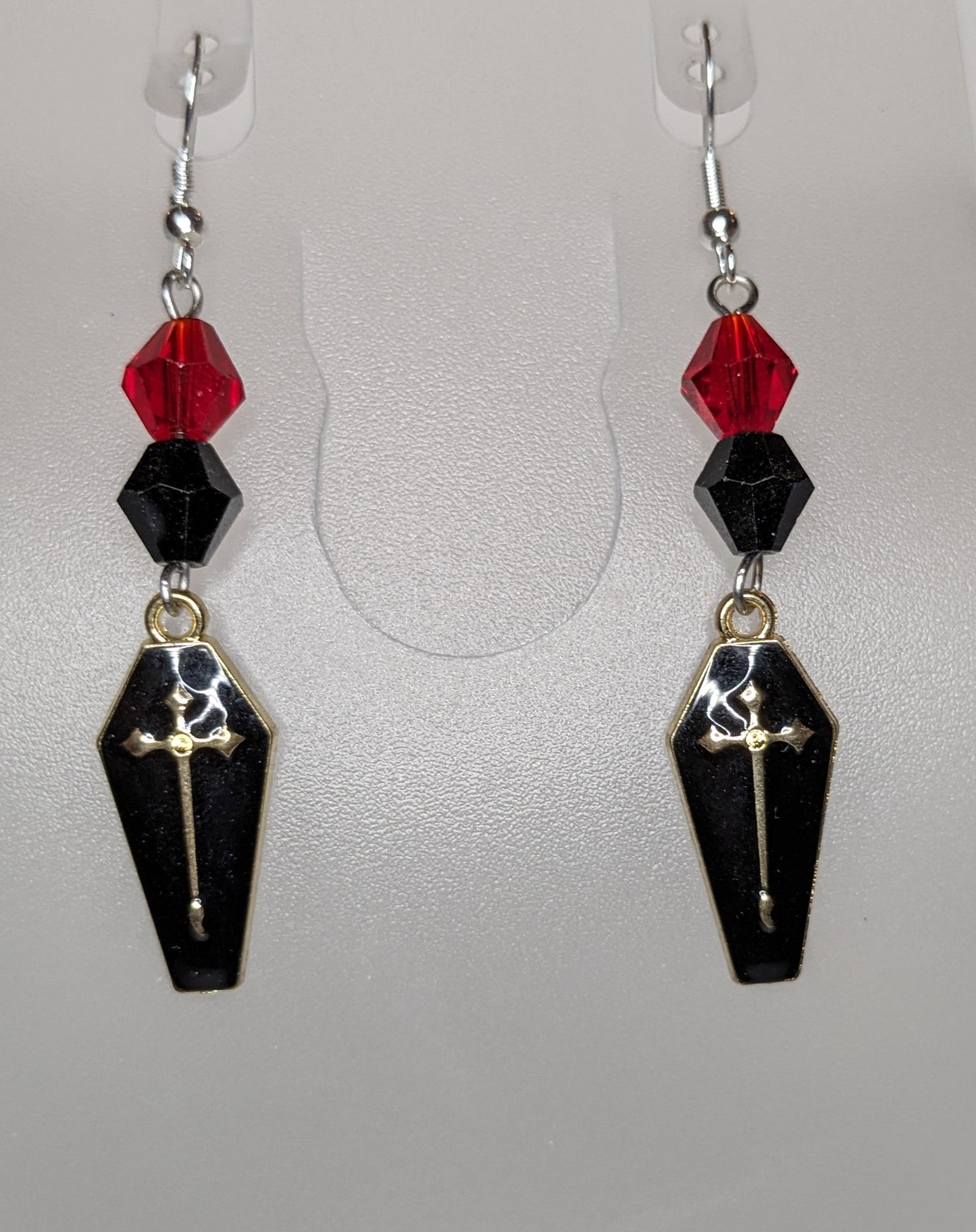 A pair of Vamp Goth Earrings featuring coffin-shaped charms enameled in black with a gold cross with faceted black and red bead accents.
