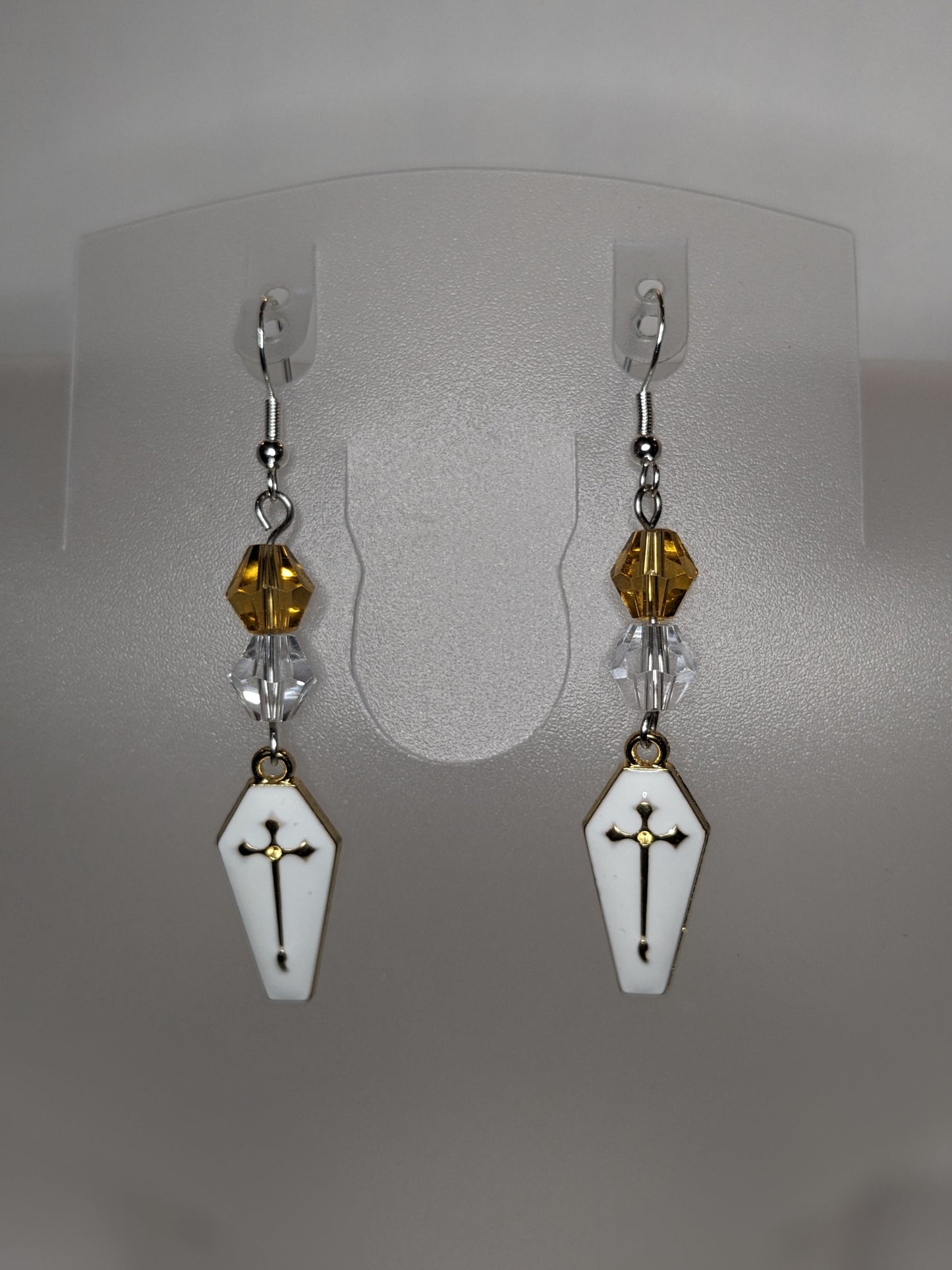 A pair of Vamp Goth Earrings featuring coffin-shaped charms enameled in white with a gold cross with faceted gold and clear bead accents.