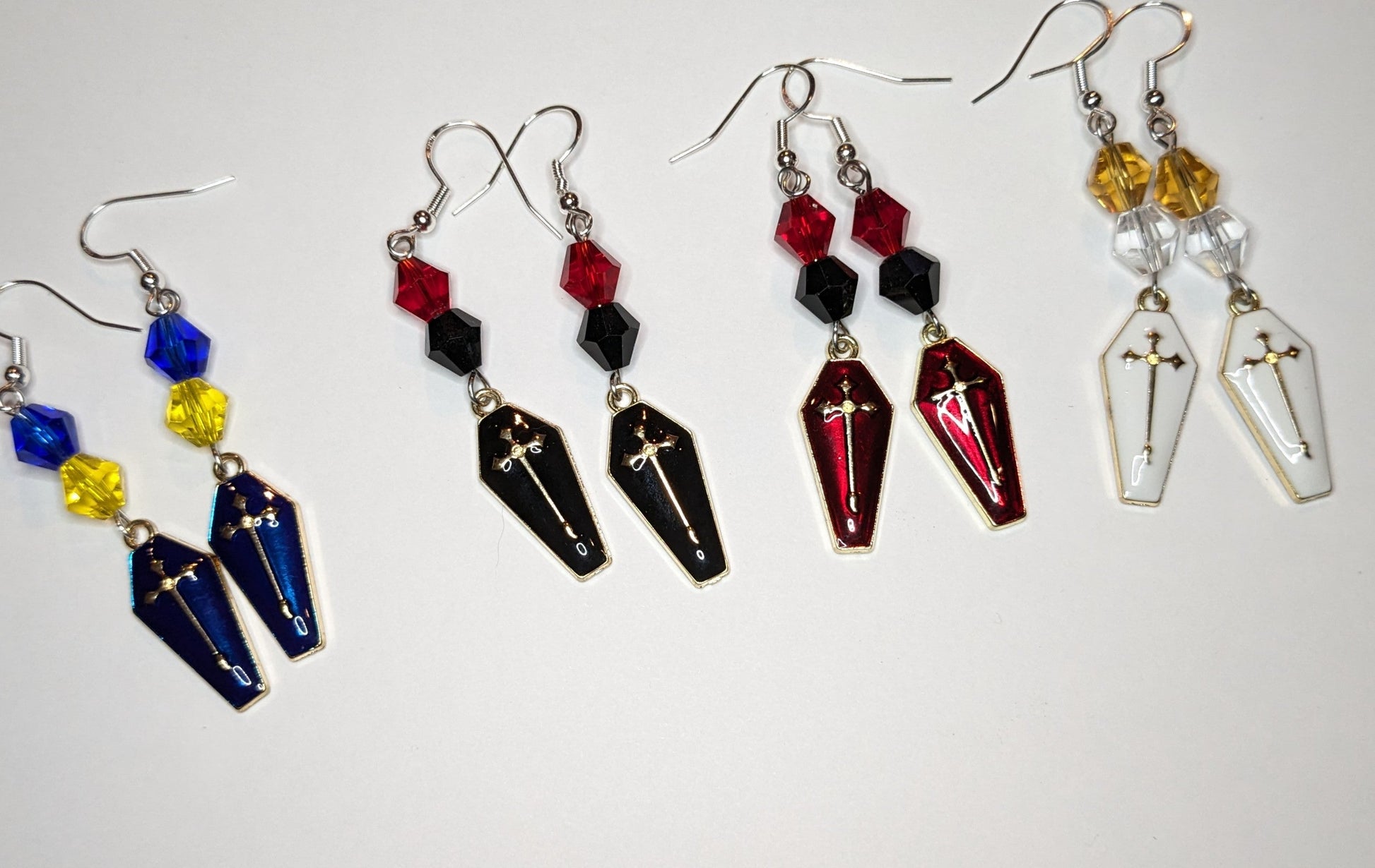 4 pairs of enameled coffin shaped earrings with bicone crystal bead accents in different colors: dark blue with gold cross and blue and yellow beads, black with gold cross and black and red beads, red with gold cross and red and black beads, and white with gold cross and gold and clear beads